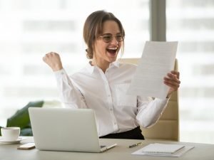 Das Bild zeigt eine überglückliche Frau mittleren Alters, die an einem stilvollen, gut organisierten Schreibtisch sitzt. Sie hält triumphierend ein Exemplar ihrer fertigen Bewerbung in den Händen, deutlich sichtbar mit einem auffälligen und professionellen Layout. Der Laptop auf dem Tisch zeigt einen aktiven Chat mit ChatGPT, wobei der Bildschirm Textausschnitte einer Bewerbung und hilfreiche Tipps von ChatGPT anzeigt. Die Frau hat eine europäische Abstammung und trägt Business-Casual-Kleidung, was eine professionelle und zugleich bequeme Atmosphäre schafft. Der Hintergrund des Raumes ist neutral und unaufdringlich, mit einem Fokus auf die Frau und ihre Tätigkeit. Das Zimmer ist hell beleuchtet, wobei das Licht eine warme und einladende Stimmung erzeugt. Auf dem Schreibtisch sind weitere Bewerbungsunterlagen und ein Smartphone zu sehen, was auf einen modernen, technologieorientierten Bewerbungsprozess hindeutet. ChatGPT, Bewerbungsschreiben, erfolgreiche Bewerbung, Jobbewerbung, Karrierefortschritt, digitale Assistenten, Bewerbungstipps, Online-Bewerbung